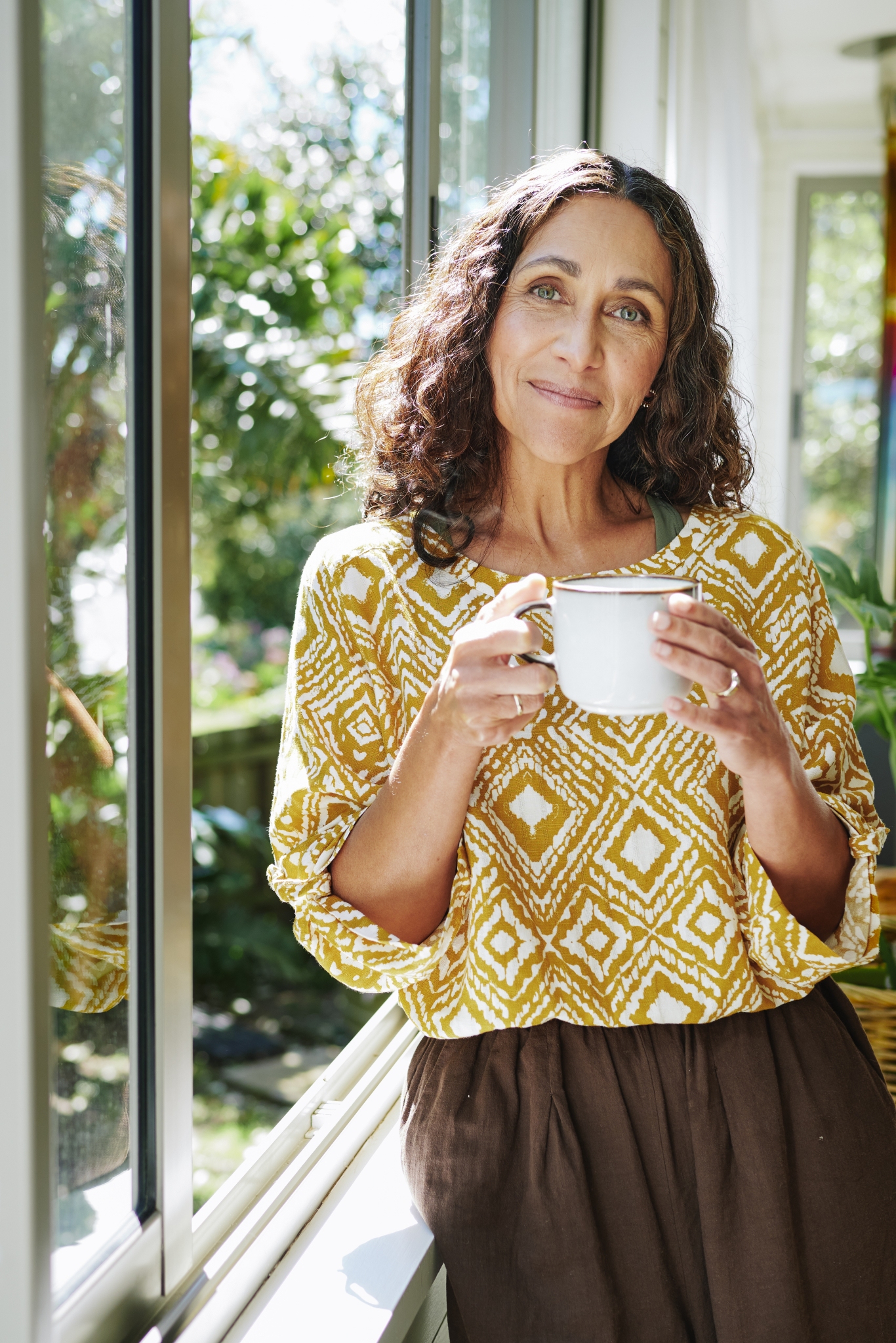 Mature woman smiling while drinking coffee by an open window at home